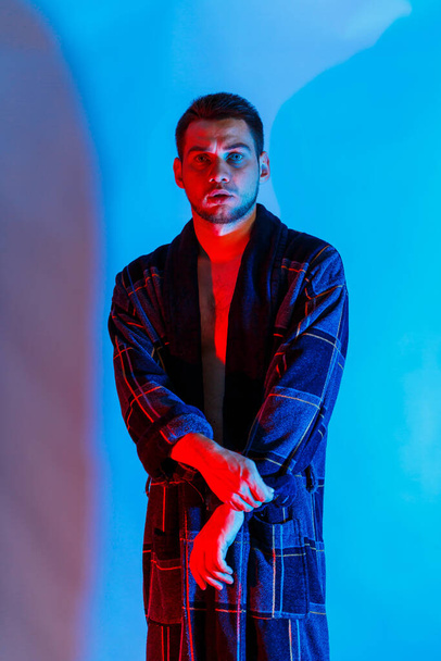 Ένας άντρας με μπουρνούζι και εσώρουχα. Στούντιο φωτισμού κόκκινο και μπλε σε ένα ανδρικό σώμα. Εξωφρενικό μοντέλο. - Φωτογραφία, εικόνα