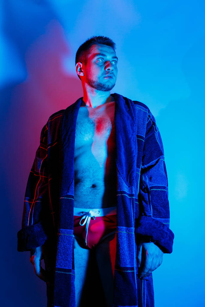 A man in a bathrobe and underwear. Studio lighting red and blue on a male body. Outrageous guy model - Photo, Image