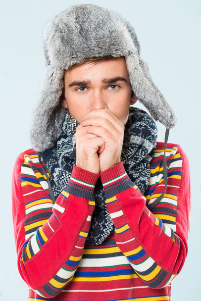 Handsome man in striped sweater, scarf and a furry hat is feeling cold over a blue background - Photo, Image