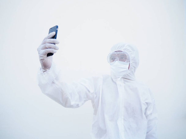 PPEスイート制服の若い医師や科学者は、家族や友人との自撮りやビデオチャットのために彼の携帯電話を使用している間。コロナウイルスまたはCOVID-19の概念分離白背景 - 写真・画像