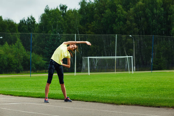 Женщина делает спортивные упражнения на открытом воздухе. Брюнетка в жёлтой спортивной рубашке и шортах на стадионе. - Фото, изображение