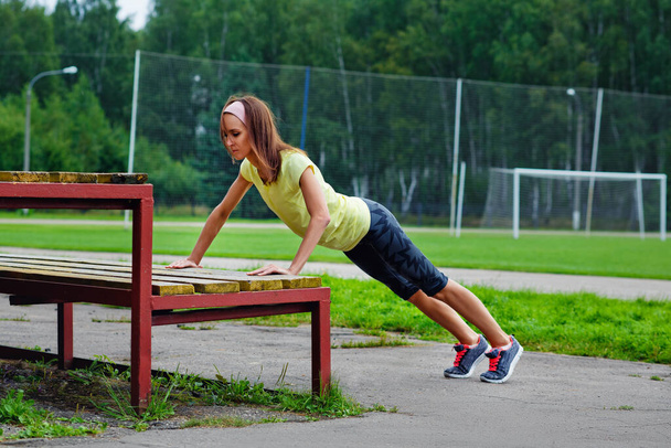 Giovane bruna sottile attraente nel fare esercizi su una panchina in uno stadio all'aperto, allenamento di base e concetto di fitness. - Foto, immagini