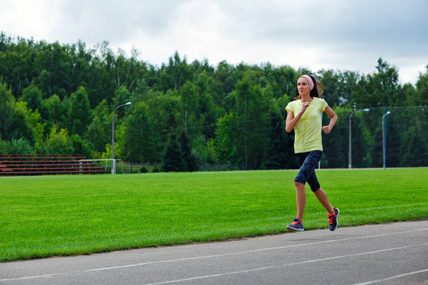Ein schönes Fitness-Mädchen in gelbem Sporttop und Leggings joggt durch das Stadion. An einem warmen Sommertag läuft sie schnell. Die Sportlerin engagiert sich in ihrer normalen Sportpraxis. - Foto, Bild
