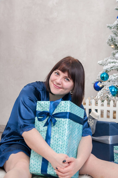 A middle-aged woman hugs a gift box near the Christmas tree at home, new year's joyous mood - Фото, зображення