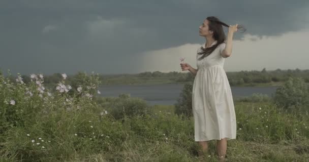 Κομψό κορίτσι χαϊδεύοντας μακριά μαλλιά για το καλοκαίρι αέρα βροχερό τοπίο χώρα φόντο. Νεαρή γυναίκα με λευκό φόρεμα στέκεται μόνη της στη φύση σε εξωτερικούς χώρους αργή κίνηση αντιγραφή χώρου. Τροποποίηση έννοιας ελευθερίας - Πλάνα, βίντεο