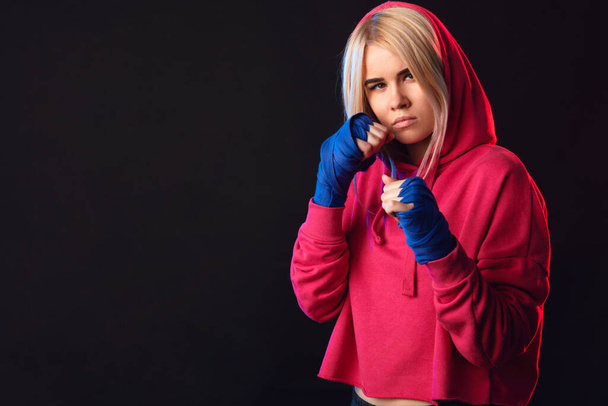 Woman in pink wear with hood on, practicing punches while standing in dark gym - Фото, изображение