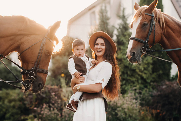 Egy anya kalapban a fiával a karjában két gyönyörű ló mellett áll a természetben. egy gyermekes családot lefényképeznek lovakkal. - Fotó, kép