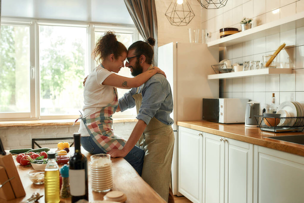 Young man looking at his girlfriend, hugging while standing in the kitchen. Couple embracing while making pizza with vegetables indoors. Cooking together, hobby, lifestyle - Фото, изображение