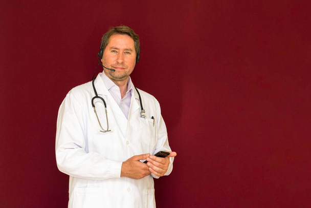 Young caucasian doctor with stethoscope talking with patient online red studio background - Photo, Image