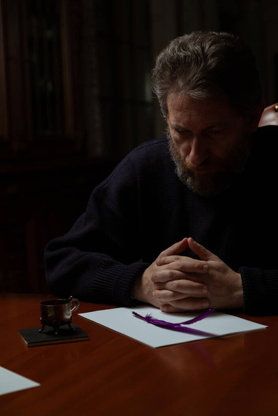 man with a beard sits at a large polished table and reflects on a white sheet. In the hand is a goose feather. Nearby is a metal inkwel - Zdjęcie, obraz