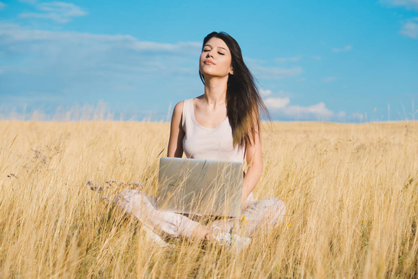 Egy fiatal, kedves nő, aki egy laptopon dolgozik sárga mezőn. Munka a természetben. - Fotó, kép