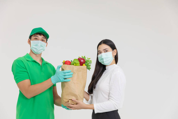 The smart food delivery service man in green uniform wearing face mask handing fresh food to recipient and young woman customer receiving order,express delivery, food delivery, online shopping concept. - Photo, Image