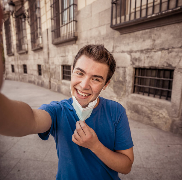 COVID-19 outbreak. Young man in city street wearing protective surgical mask and using mobile phone video calling friends and family. Stay connected in the New Normal and mandatory use of face mask. - Photo, image