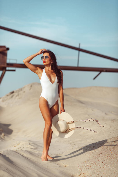 A young beautiful girl with a good figure, around only white sand and blue sky, and on her a white swimsuit and beautiful accessories - Fotoğraf, Görsel