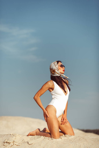 A young beautiful girl with a good figure, around only white sand and blue sky, and on her a white swimsuit and beautiful accessories - Foto, immagini