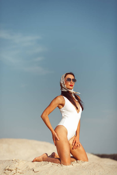 A young beautiful girl with a good figure, around only white sand and blue sky, and on her a white swimsuit and beautiful accessories - Foto, Bild