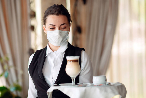 A female Waiter of European appearance in a medical mask serves Latte coffee. - Фото, зображення