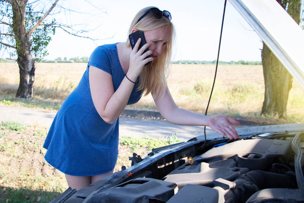 Блондинка смотрит на двигатель, который не работает. Женщина пытается объяснить механику причину срыва с помощью смартфона и позвать на помощь. Концепция разбивки дорог - Фото, изображение