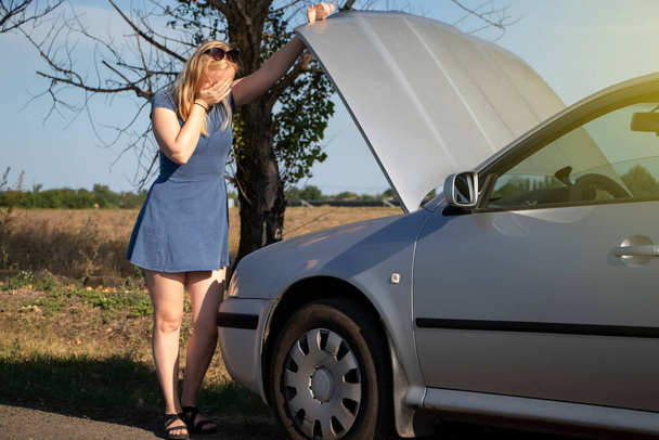 A blonde in a blue sundress votes on the road, near her car. An open bonnet indicates a breakdown in the vehicle. The concept of helping motorists in a difficult situation. Sweet girl in trouble - Photo, Image