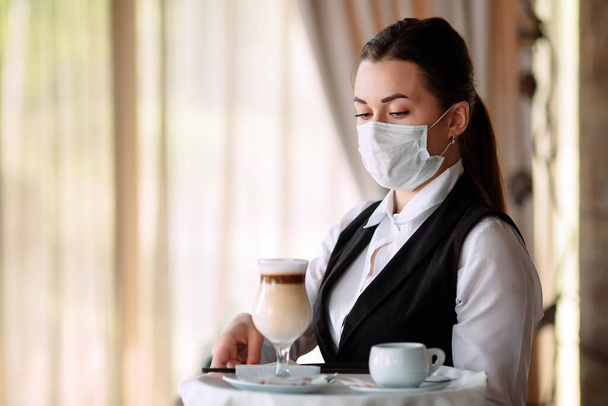 A female Waiter of European appearance in a medical mask serves Latte coffee. - 写真・画像