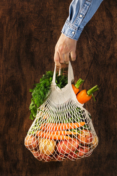 Mesh bag with vegetables and herbs in female hand. Woman hold string net shopping bag on wooden background. Modern reusable shopping, eco friendly lifestyle, zero waste concept - Foto, Bild