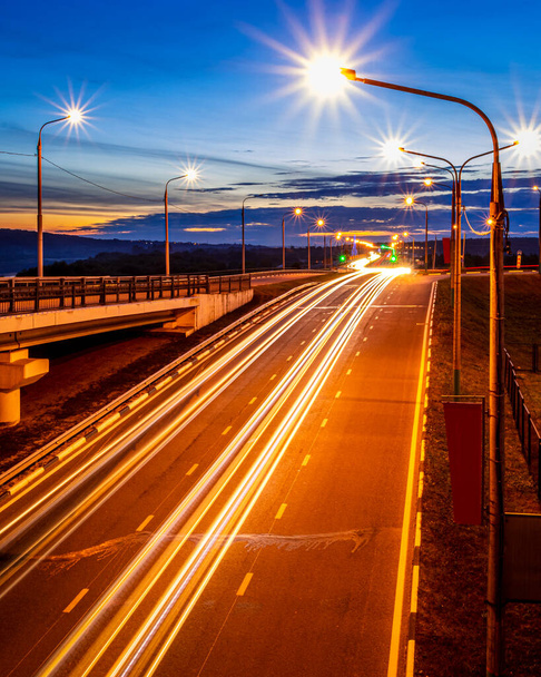Ślady reflektorów z samochodów poruszających się nocą po moście, oświetlone latarniami. Streszczenie krajobraz miasta z autostradą o zmierzchu. - Zdjęcie, obraz