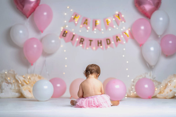 First year's birthday.a happy little girl in pink tutu skirt sits in background with garlands and balloons on a background with garlands and pink balloons, celebrating her first birthday. Copy space. - Fotografie, Obrázek