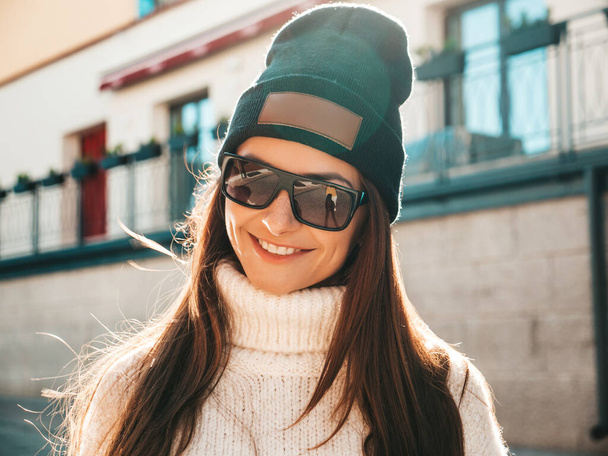 Portrait of beautiful smiling model. Female dressed in warm hipster white sweater and beanie. Trendy woman posing in the street at sunset in sunglasses - Foto, afbeelding