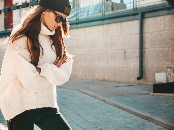 Portrait of beautiful smiling model. Female dressed in warm hipster white sweater and beanie. Trendy woman posing in the street at sunset in sunglasses - Foto, Imagem