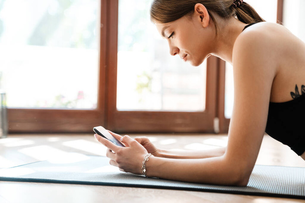Fit young sports woman using cellphone while doing exercise on a fitness mat at home. - 写真・画像