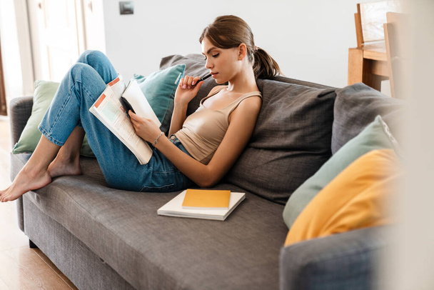 Attractive young woman studying while sitting on couch at home, taking notes - Фото, зображення