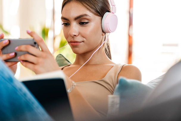Beautiful young woman listening to music with headphones, writing in a diary on a couch, using mobile phone - Foto, Imagem