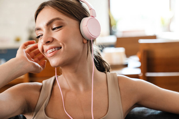 Attractive cheerful young woman listening to music with headphones, sitting on a couch - Photo, image