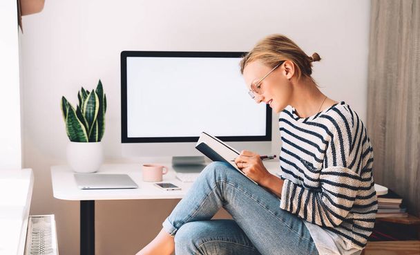 Woman freelancer working at home office. Girl wearing in glasses works with new startup project. Female thinking and writing in planner or diary at workplace of minimalist style with computer - Φωτογραφία, εικόνα