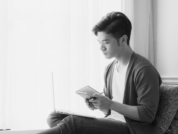 Молодой азиат пишет в книге при использовании ноутбука - Фото, изображение