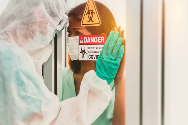 Preoccupazioni tra le donne infette Coronavirus (Covid-19) e il giovane che indossa indumenti protettivi PPE. Guardando insieme attraverso il vetro nella stanza di contenimento e monitorando attentamente il trattamento - Foto, immagini