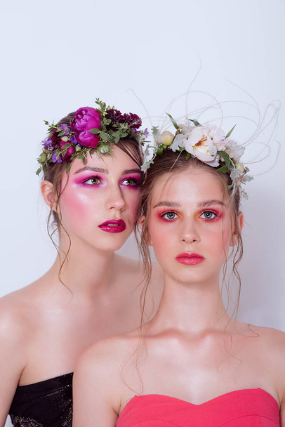 Two fashion beauty models sister girls with professional bright make-up, a hoop of fresh spring flowers on their heads, with red lilac lipstick are posing against a gray uniform background. Clean skin - Photo, Image