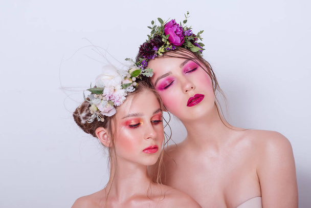 Two fashion beauty models sister girls with professional bright make-up, a hoop of fresh spring flowers on their heads, with red lilac lipstick are posing against a gray uniform background. Clean skin - Photo, Image