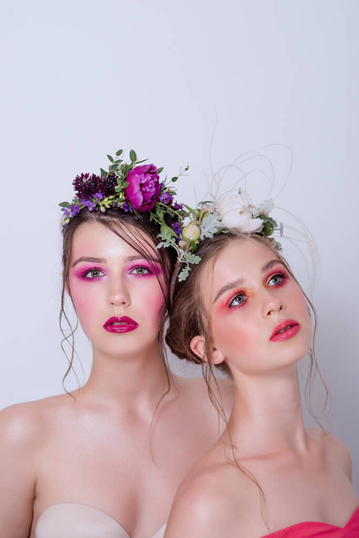 Two fashion beauty models sister girls with professional bright make-up, a hoop of fresh spring flowers on their heads, with red lilac lipstick are posing against a gray uniform background. Clean skin - Zdjęcie, obraz