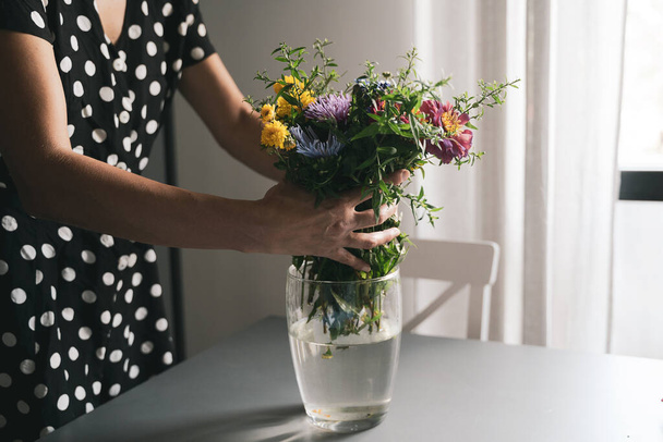 Siyah elbiseli, beyaz benekli genç beyaz bir kadın, bir buket taze çiçek hazırlıyor. Gri bir masanın üzerindeki cam vazoda, gün ışığında. - Fotoğraf, Görsel