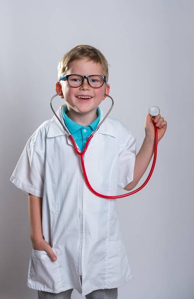 Хлопчик грає з лікарем зі стетоскопом. Дитина в окулярах ізольована над білим. Палець вгору. Білий одяг
. - Фото, зображення