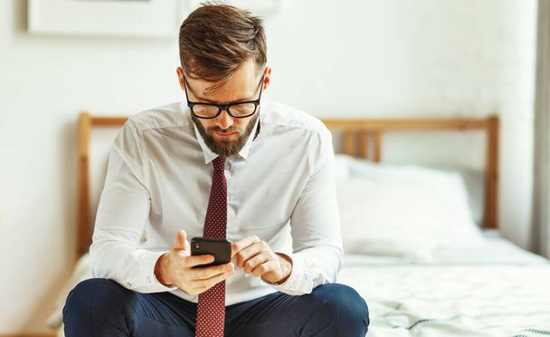 Ernsthafter junger bärtiger Geschäftsmann in formalem Outfit und Brille, der auf dem Bett sitzt und Nachrichten auf dem Handy sendet, während er Geschäftsprobleme von hom aus löst - Foto, Bild
