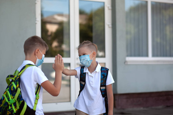 Schoolkinderen, met maskers en rugzakken, begroeten veilig voor de deur van de school. Terug naar school. studenten zijn klaar voor nieuw schooljaar. - Foto, afbeelding