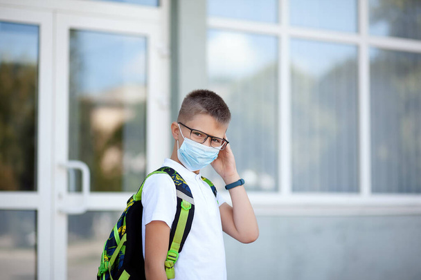 Возвращайся в школу. Подросток в очках в маске и рюкзаках защищает и обезопасит от коронавируса. Ребенок ходит в школу после пандемии. Стоя рядом со школой. студенты готовы к новому году. - Фото, изображение