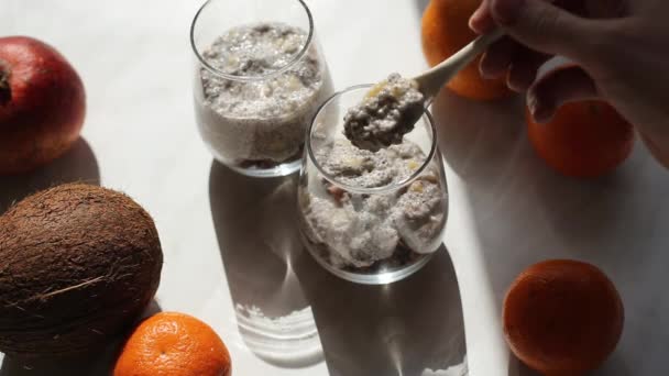 Der Herstellungsprozess von Pudding aus Chiasamen mit Kokosmilch und Müsli. Chia-Samen ins Glas geben. Gesunde vegane Ernährung. Sonnenhartes Licht in der Küche aus dem Fenster. - Filmmaterial, Video