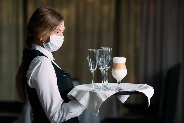 A female Waiter of European appearance in a medical mask serves Latte coffee. - Photo, image