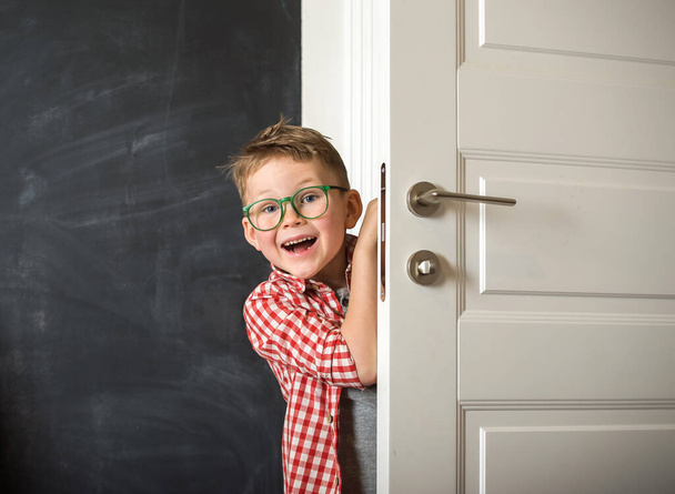 Uśmiechnięty, młody chłopiec podglądający przez drzwi. Dzieciak wrócił ze szkoły. Szczęśliwe dziecko w okularach. Ręka na rączkę. - Zdjęcie, obraz