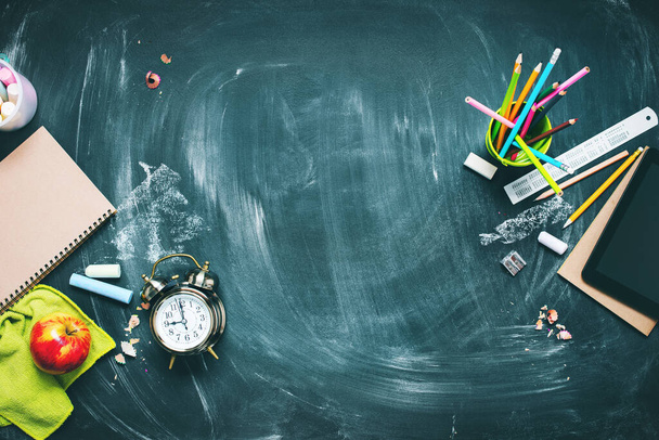 Back to school concept. Office supplies. Stationery on green blackboard background alarm clock, color chalks, pencil, apple, notebook. Copy space. Top view. Flat lay. - 写真・画像