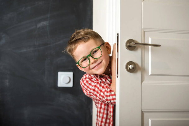 Un niño sonriente asomándose por la puerta. El chico de la escuela vuelve de la escuela. Niño feliz con gafas. Mano a mano. Sorpresa.. - Foto, Imagen