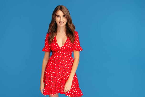 Charming young woman with bright smile in red dress posing over blue background. - Zdjęcie, obraz